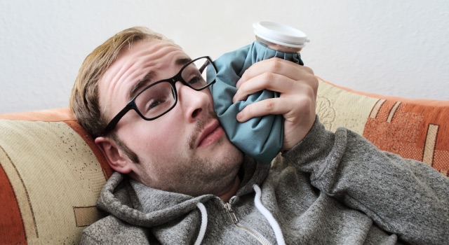 close-up of man with glasses and light hair laying on a couch holding an ice pack against his face