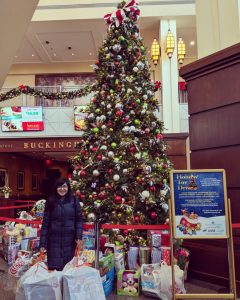 A woman with glasses standing in front of a Christmas toy drive, smiling and holding bags of toys